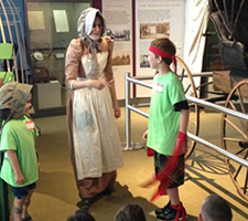 An educator in historical dress speaks to two children.