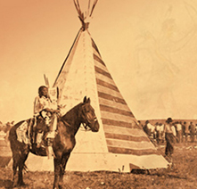 A Native American man sits on a horse with a tipi and people visible in the background