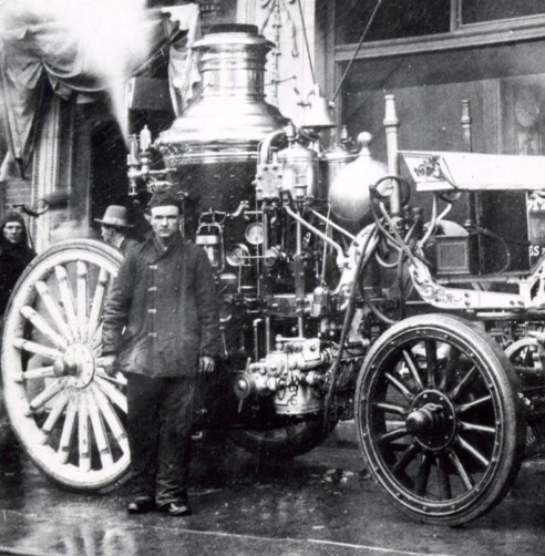 A man stands in front of an elaborate steam pumper fire engine in this black-and-white historical photograph. 