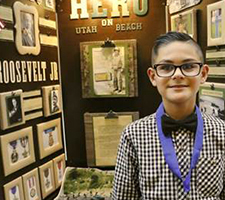 A young man in glasses stands in front of his History Day exhibit.
