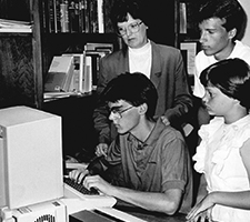A young man uses a 1990s-era computer  while others look on.