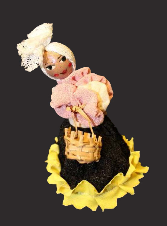 A small doll with a painted wooden face wears a headwrap and carries a tiny basket.