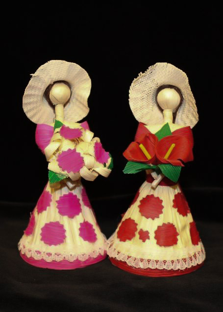 Two cornhusk dolls with hats and flowered dresses holding flowers in their arms.