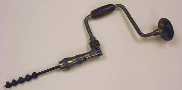 The drill has spiral bit, a U-shaped crank handle, and a wooden head. One hand turns the crank while the other holds the wooden head to steady it.