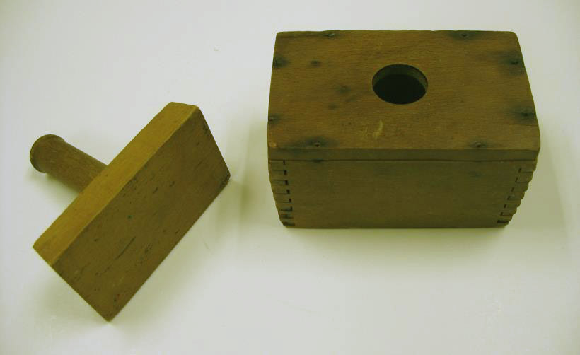 A wooden box with a hole on one side, and a rectangular piece of wood used to put butter through the hole.