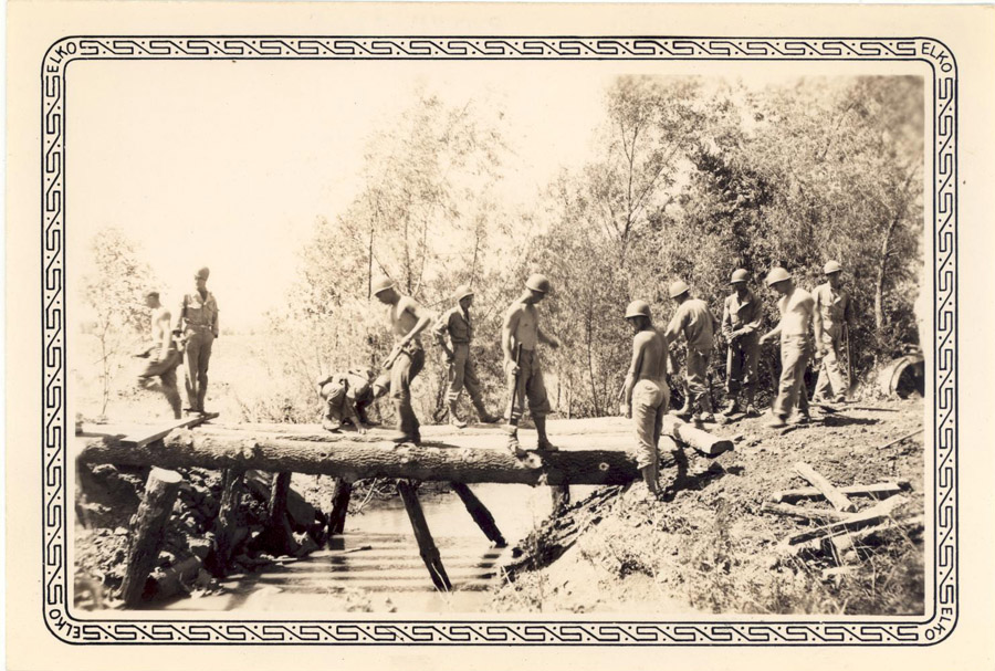 African American soldiers stand and work on a log bridge over a stream in a forested area. A few soldiers stand to the side and observe. Bridge supplies are strewn around the grass.
