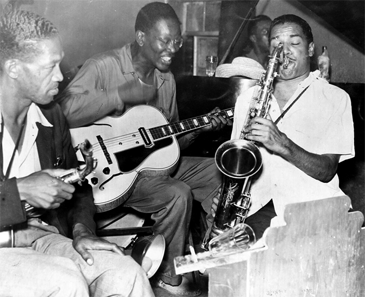 Three Black men sit close together playing instruments including the guitar and saxophone. 