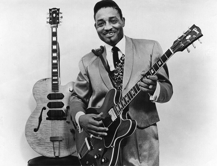 A picture of a Black man in a suit holding a guitar and standing in front of another guitar. 