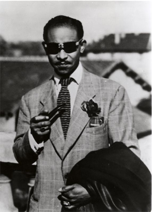 A Black man wearing a suit and sunglasses and holding a coat. 