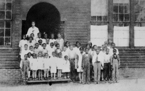 A picture of a group of African American schoolchildren standing outside of a school entrance. The teacher stands in the back. 