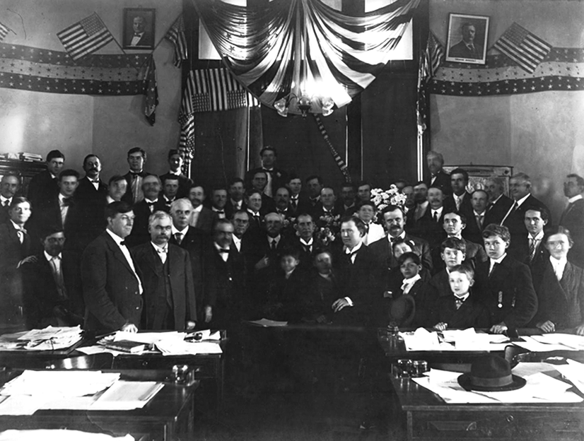 A group of about forty white men in suits with about seven children standing behind desks and in front of a wall decorated with patriotic decorations. 