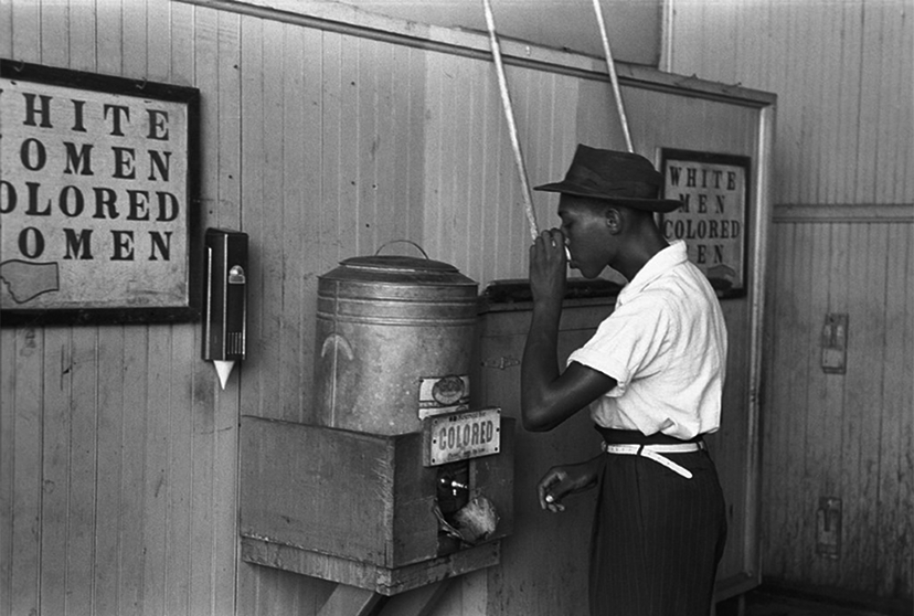 A young Black man drinks from a water container labeled, 'Colored' in front of other signs that read: 'White Women, Colored Women' and 'White Men, Colored Men.'