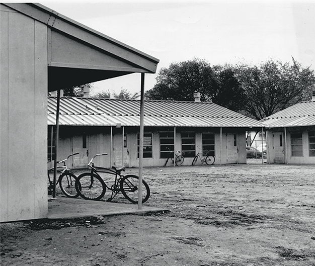 Buildings with metal roofs built in a U shape. Bicyles are parked in front of two of the buildings. The lot is bare dirt. 