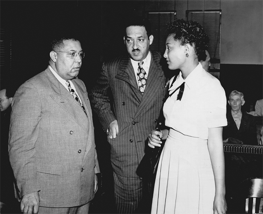 Two Black men in suits speak with a Black woman wearing a dress. They are inside a court room. 
