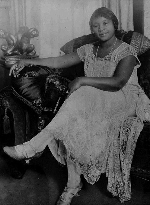 An African American woman sitting in a chair with one leg over the other, and smiling at the camera.