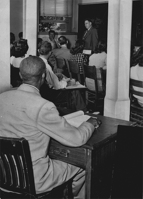 A man teaches in front of about thirteen white students sitting at desks. A Black man sits at the back, separate from the others. 