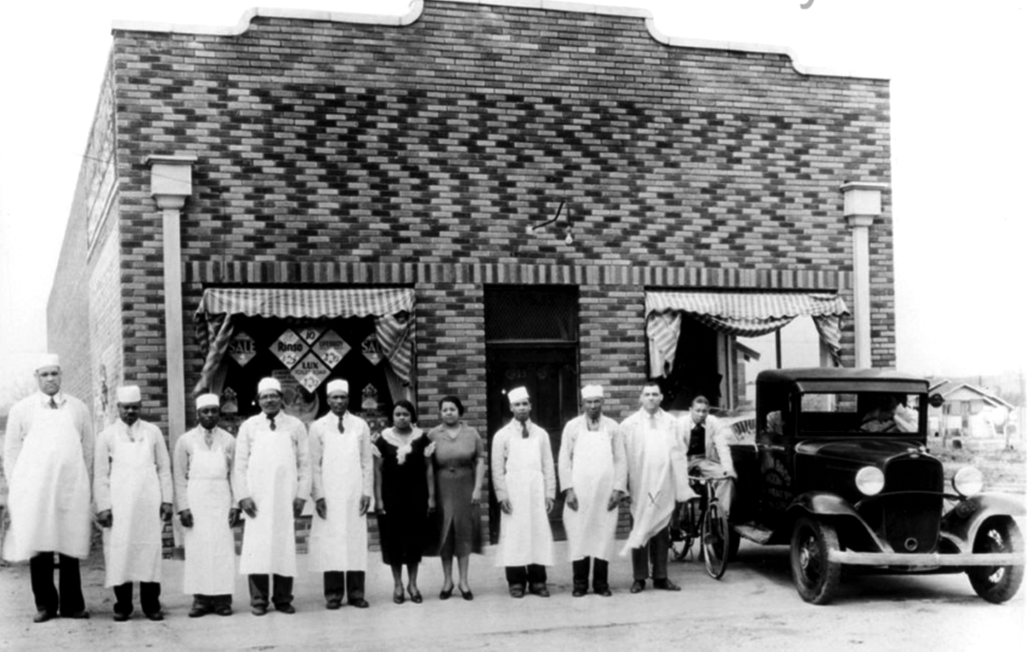 A group of people standing outside a brick building next to a car. Most are wearing long aprons and hats. 