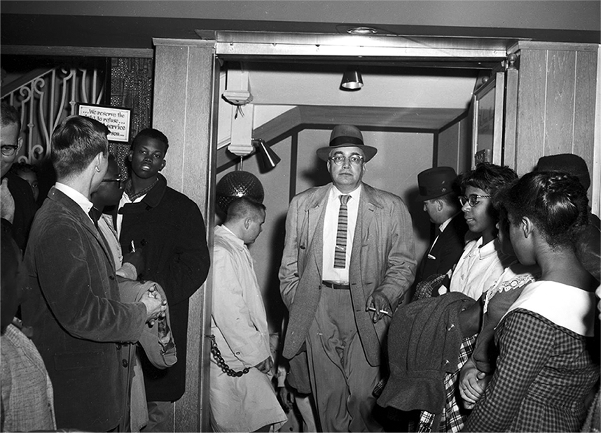 An older Black man in a suit, coat, and hat stands in the middle of a group of younger people inside a hallway. 
