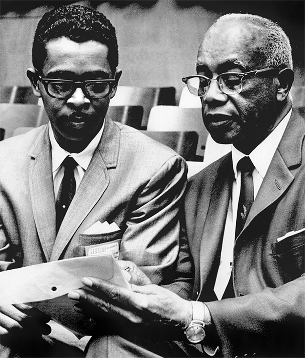 Two Black men in suits, one older, look over a document. 