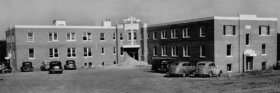 A two-story building with two wings. Several cars are parked in front of it. 