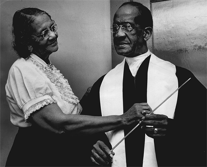An older Black man in a choir robe holds a conductor’s baton while his wife  adjusts the robe.  