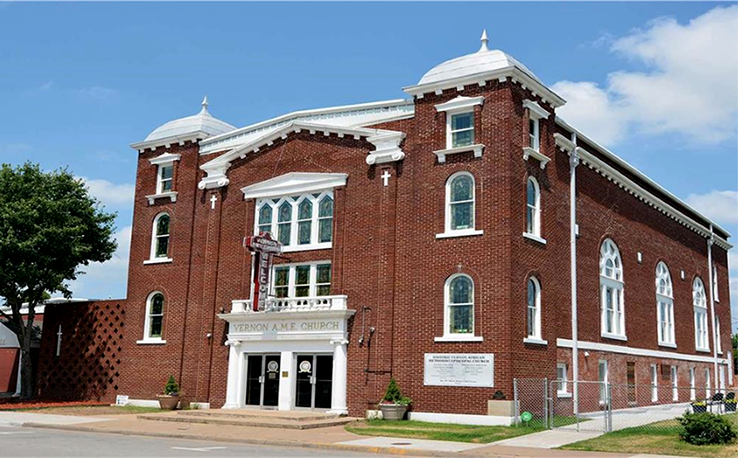 A large, multi-story red brick building with white decoration and a sign reading Vernon AME Church. 