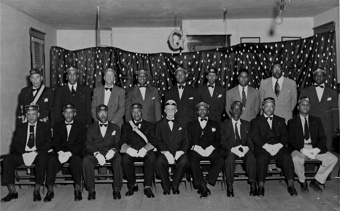 A group photo of eighteen Black men, half standing, half sitting. Most are wearing white gloves and regalia. 