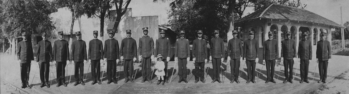 A panoramic shot of 18 Black men in uniform, holding canes. There is a small Black child in front of one of the men in the center. 