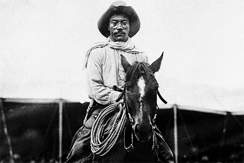 An African American man in a hat, riding a horse, looking directly at the camera.