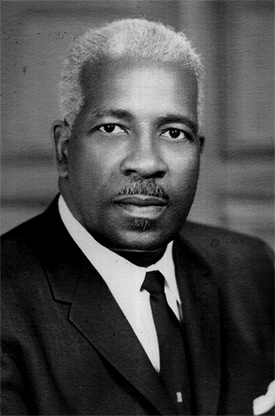 A Black man with gray hair and moustache in a black suit. 