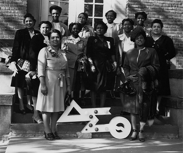 A group of twelve Black women standing in front of a building and behind a sign with greek lettering. 