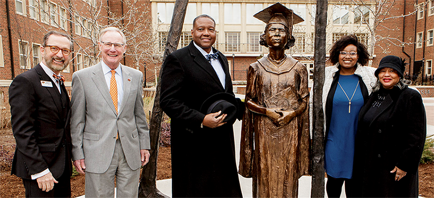 A group of people around a statue of a woman in graduation robes, hat, and holding a diploma. 