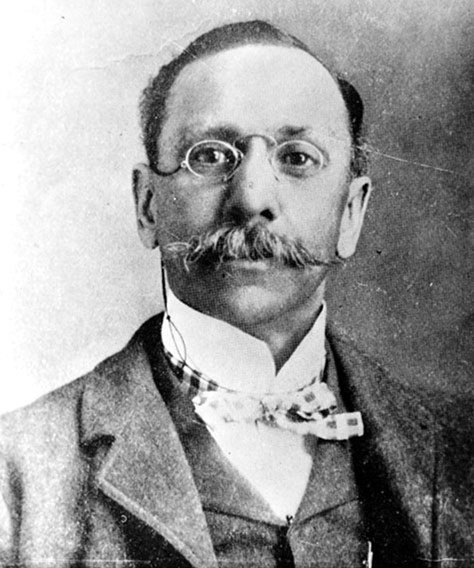 A Black man with a very large mustache, glasses, and a suit with a decorative tie. 