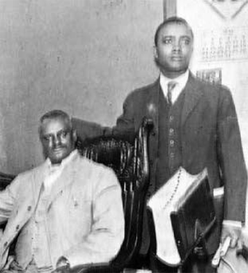A picture of two African American men, one sitting in a chair on the left and the other standing on the right.