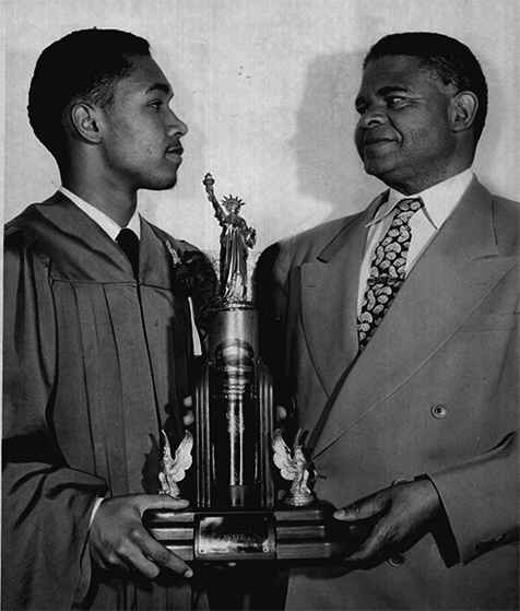 Two Black men, one older, one younger wearing graduation robe, hold a large trophy together. 