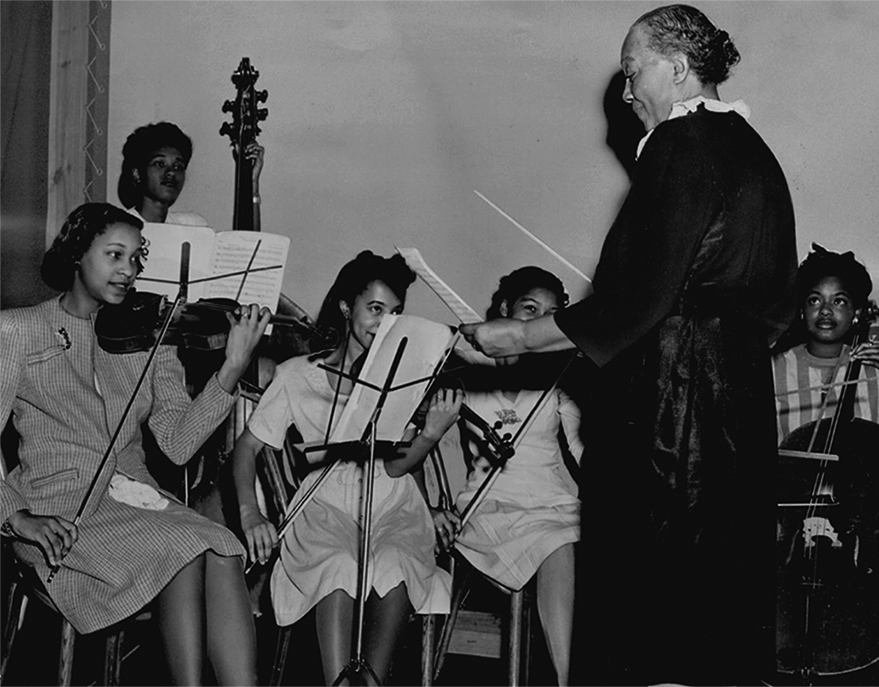 An older Black woman conducts a small group of Black women playing stringed instruments. 