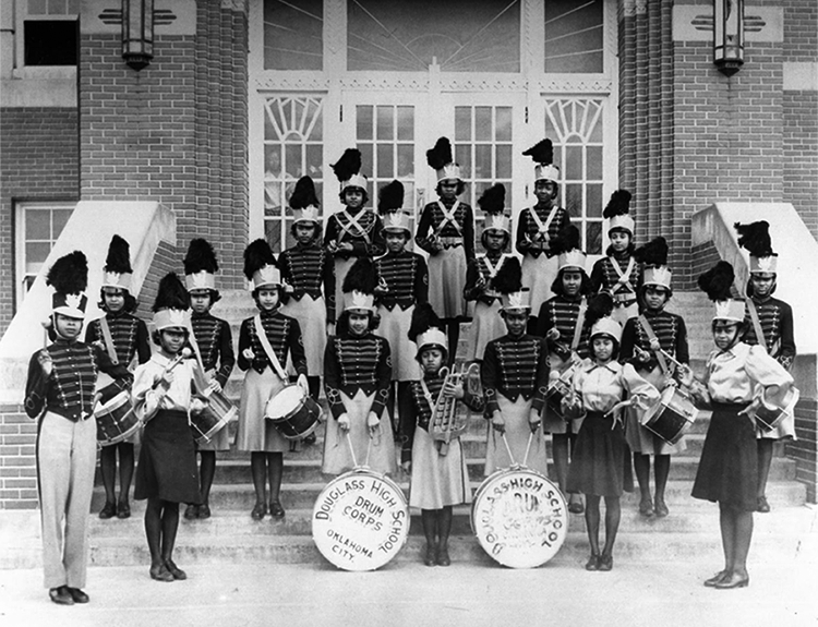 About twenty young Black women wearing band uniforms and holding instruments on the steps of a building. 