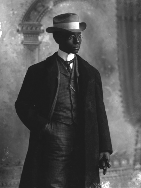 A young man wearing a suit, jacket, and hat, holding a glove. 