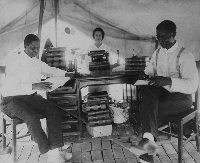 Black and white photo of two Black men and a white woman under a large tent. All three are sitting at a wood desk covered with stacks of books; the woman is typing on a typewriter.
