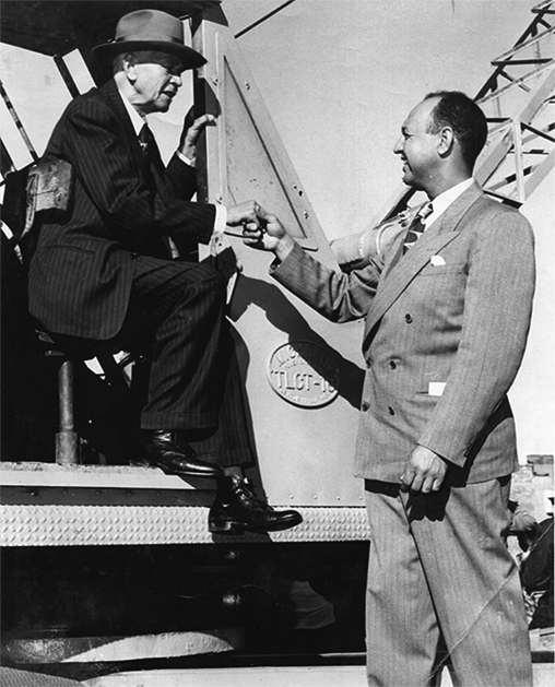 Two Black men in suits. One is sitting in a crane, the other is standing just outside the cab holding the other man's hand. 