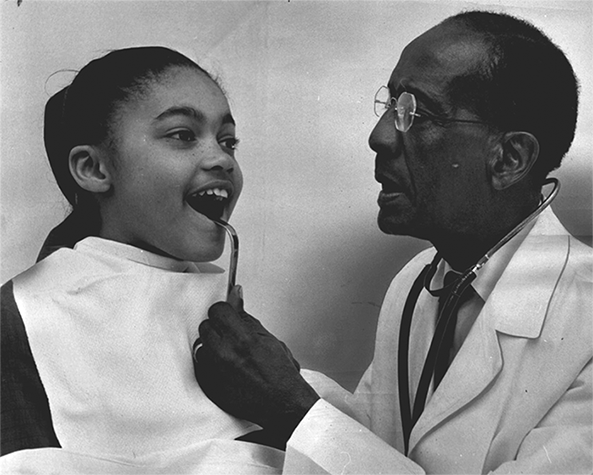 An older Black man wearing a lab coat and a stethoscope uses an instrument to examine a young girl's mouth. 