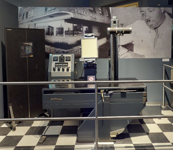 An older X-ray machine on a checkboard floor with old photos behind it. 