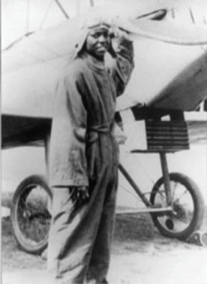 A young Black man in coveralls and aviator cap stands in front of a prop plane. 