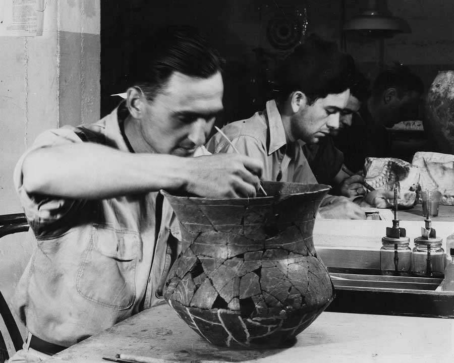 Men sit at a table inspecting a large pot and other findings from Spiro Mounds.
