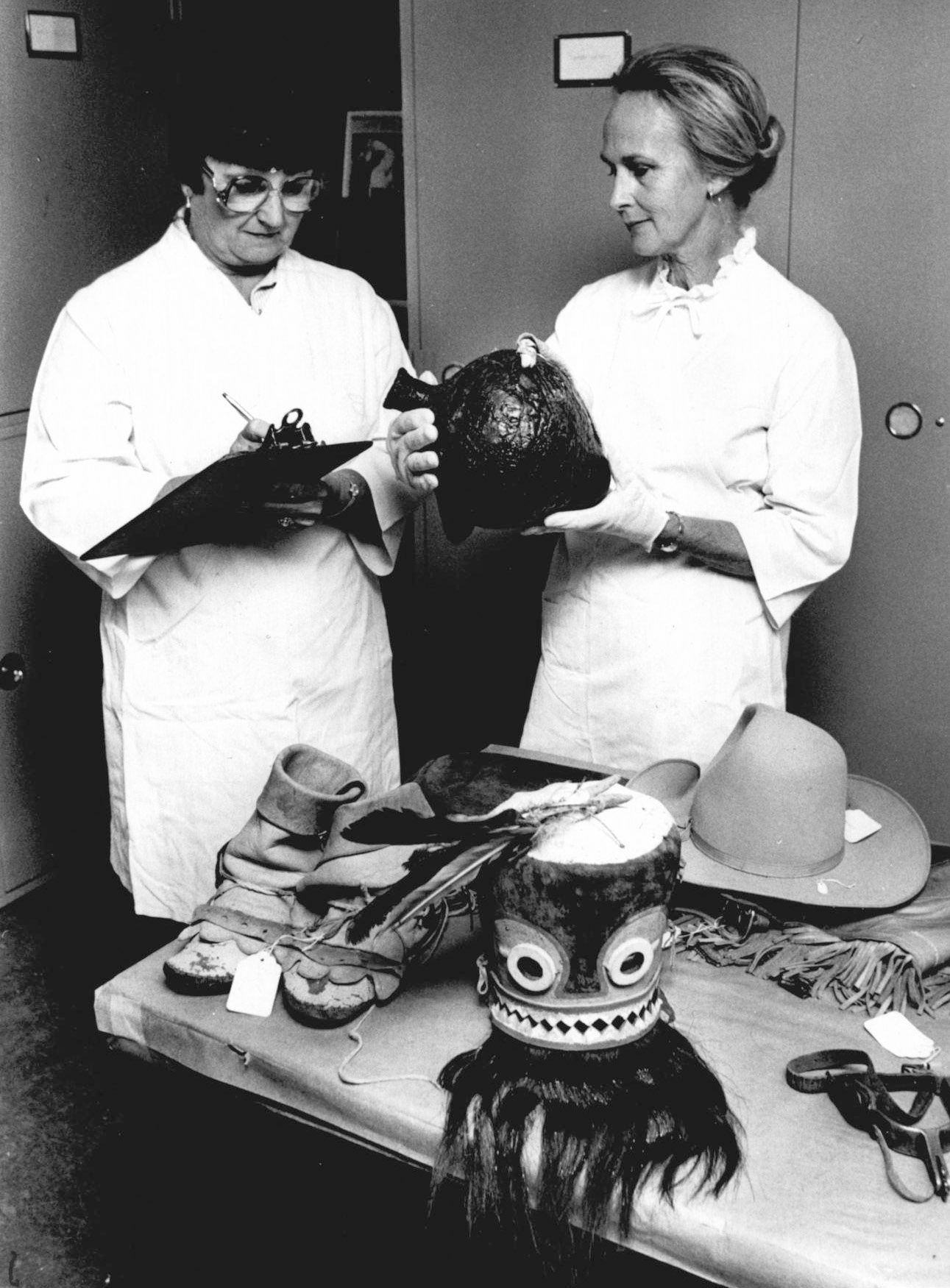 Two women stand in front of a table with many objects such as shoes, stirrups, and hats. One woman marks on a clipboard while the other holds a jar.