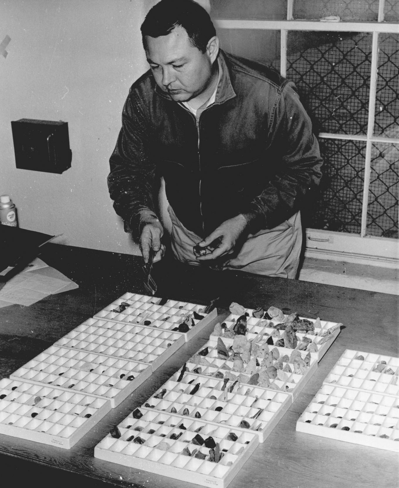 A mean  leans over a table with ten trays filled with military buttons. He is pointing at a button with a tool that he holds in his right hand.  
