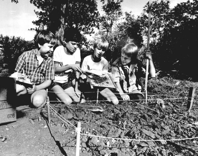 Four young boys kneel in front of a small hole blocked off by a rope tied to stakes. One holds a sifter, another holds a trowel. The boys look for artifacts in the soil using the sifter. 