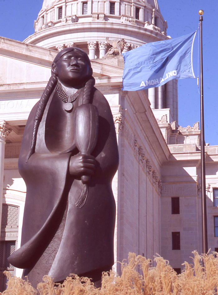 A stylized statue of a Native women stands holding a feather in her clasped hands. She wears a blanket that blows in the wind, a beaded necklace, and her hair is in long plaits. The capitol and Oklahoma flag are in the background.