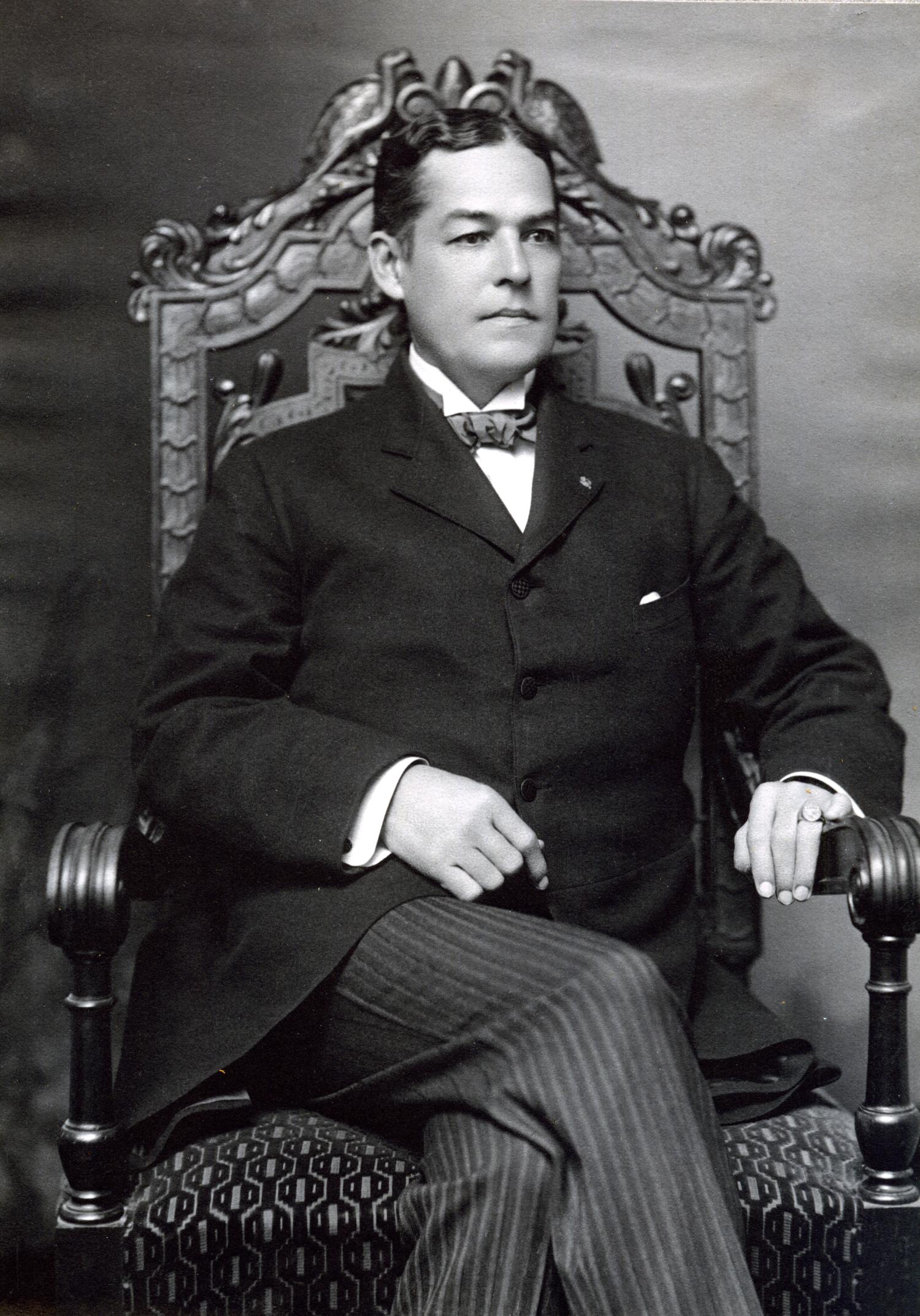 Owen wearing a formal suit, sitting in an ornate chair. He looks off to the side rather than at the camera.