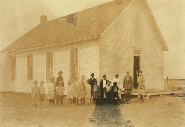 A small group of students and a few adults stand in front of a wooden schoolhouse with large windows. There are no other structures visible. 